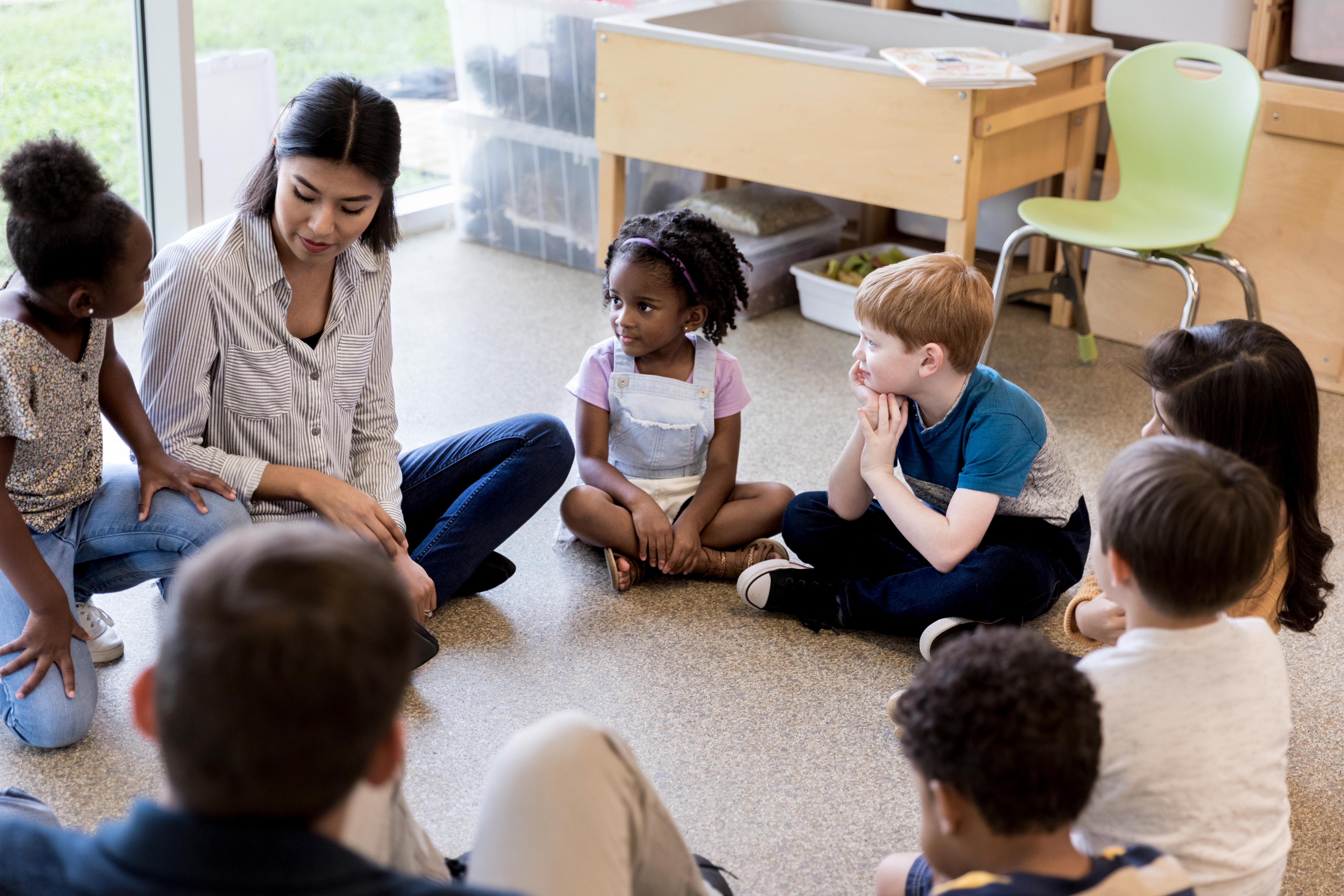 Children sit on the floor, listening to the teacher