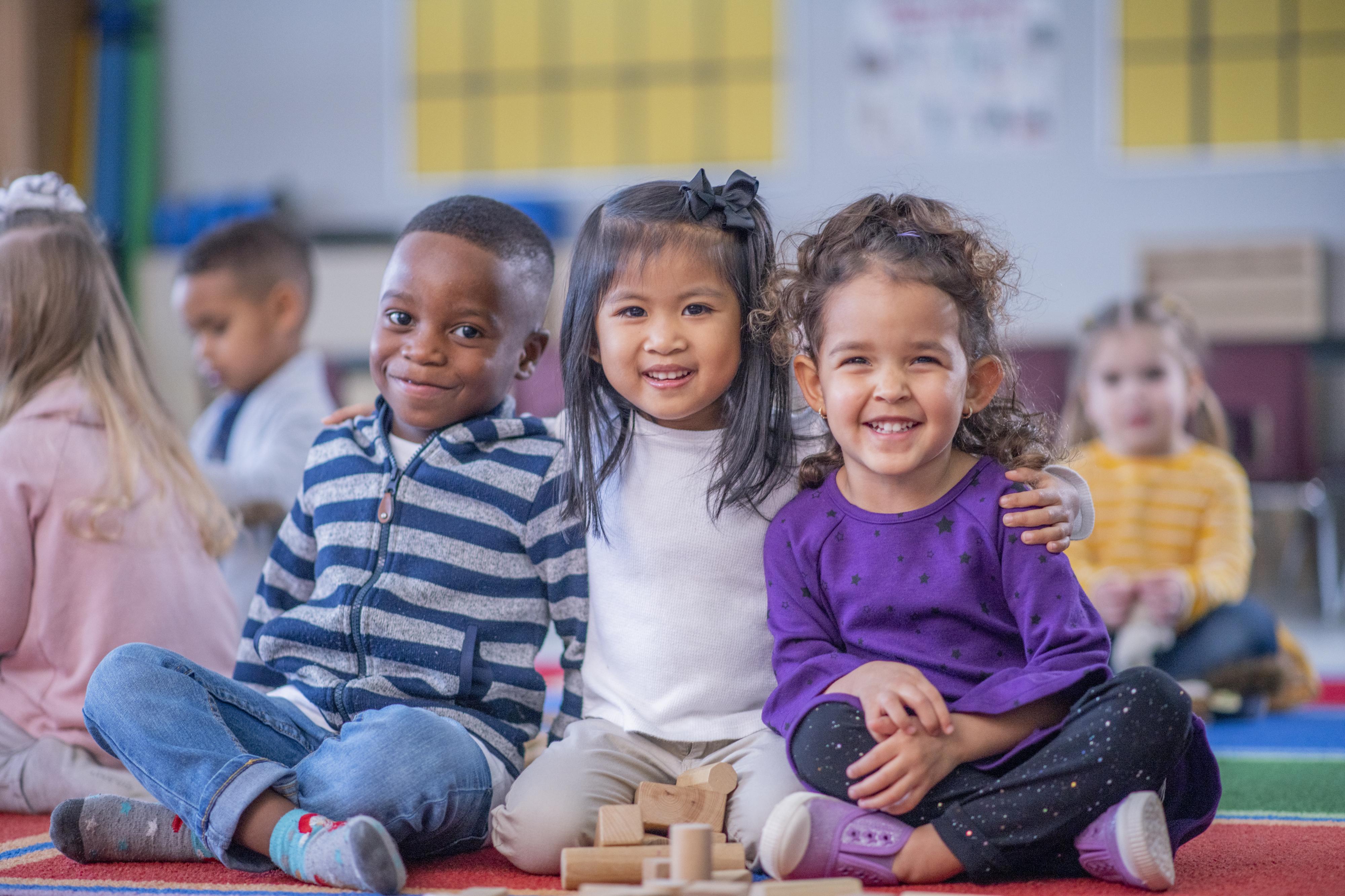 Three children sit together on the floor of a classroom and smile for the camera.