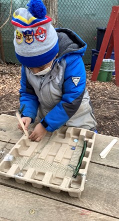 A child inventing a device from wired mesh, tongue depressors, and a cardboard tray outside