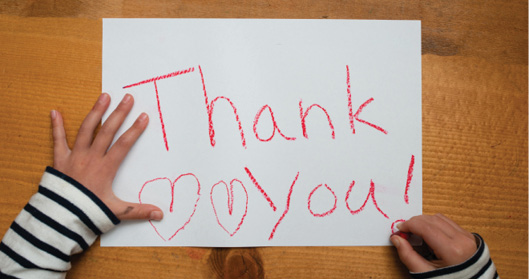 Two child's hands drawing "Thank You!" with two hearts on a white piece of paper with a red crayon