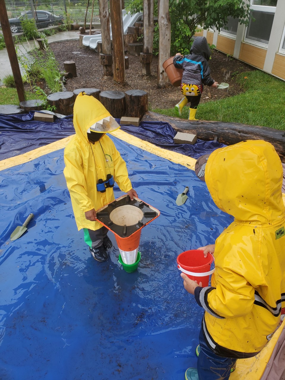 Two children in raincoats and boots play with an upside-down traffic cone outside and buckets