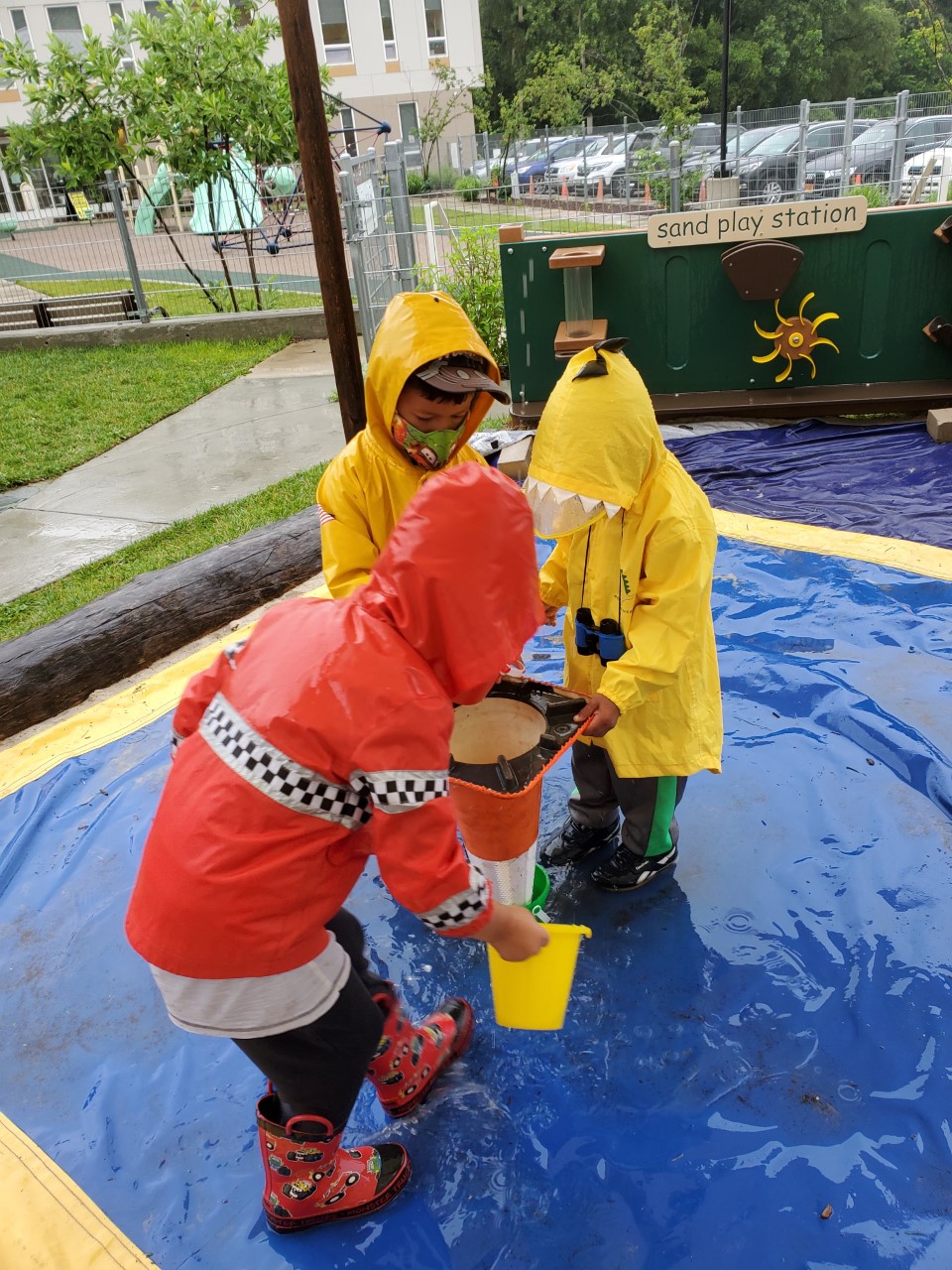 Three children in raincoats and boots play with buckets, water, and an upside down traffic cone