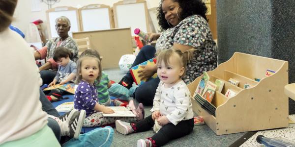 Toddlers in a classroom with their teachers, playing on the floor.