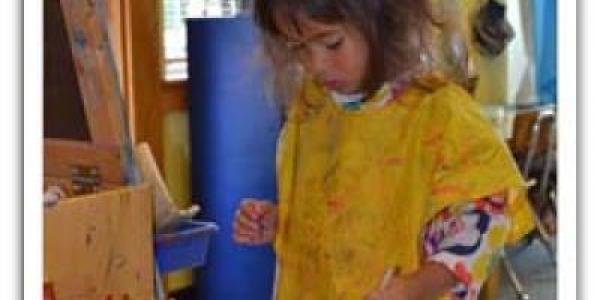 Child with paint smock on, standing near easel and paint containers.