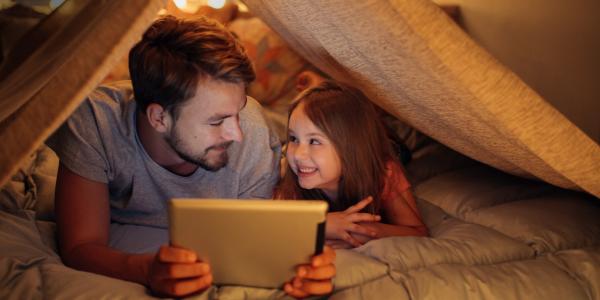 An adult reads to a child from a tablet screen under a tent indoors.