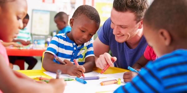 Male teacher sitting at a table with preschool children as they color