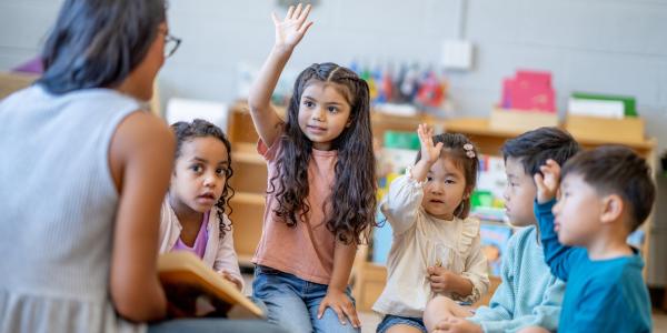a group of children listening to their teacher talking, one is raising their hand