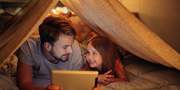 father and daughter playing on a tablet under a homemade fort. 