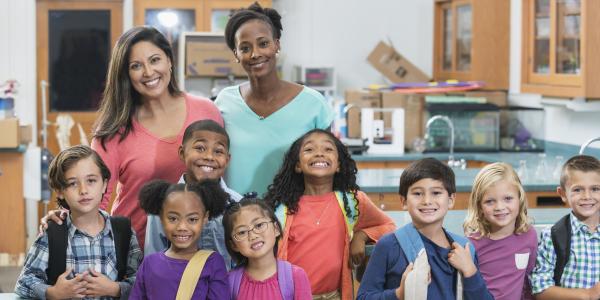 Two teachers and a group of young children standing and smiling in the classroom.