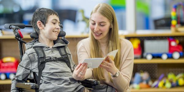 Teacher in a classroom with a young boy in a wheeler reading a card