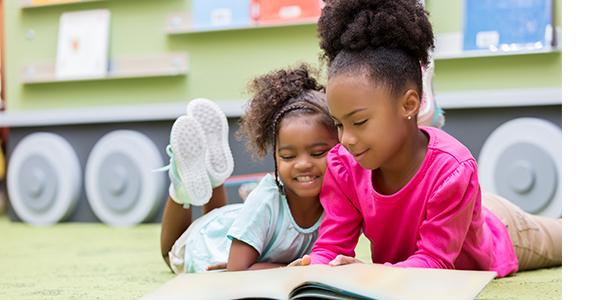 Two preschool girls on the floor in the classroom reading a book together