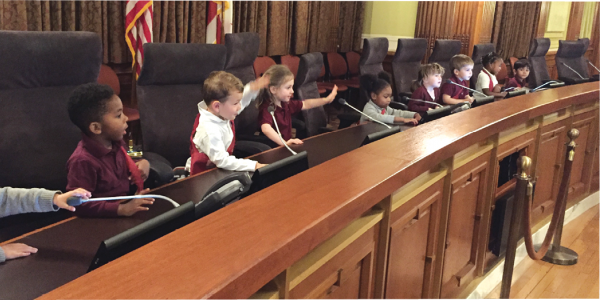 A group of preschool children sits at a city hall desk.