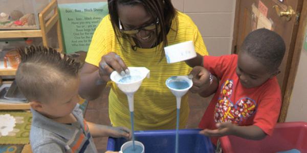 Teacher and children play with funnels at sand table.