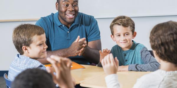 Young black male teacher of color sites at a table with group of white boys.