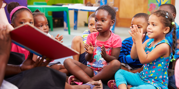 A group of children listens as a teacher reads them a book.