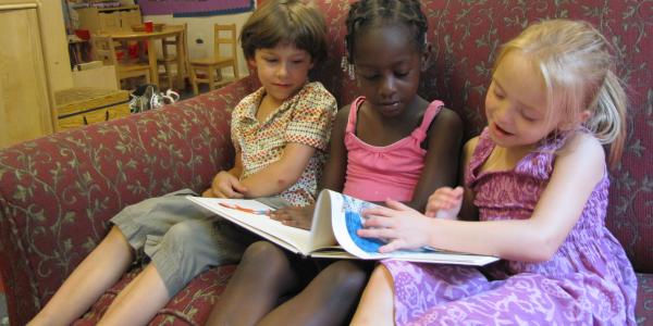 three preschoolers in a classroom sitting on a chair reading a book