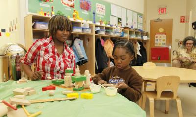 An African-American educator interacts with a young African-American girl at a low table with blocks.