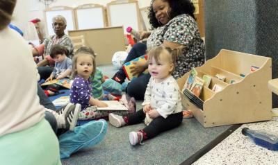 Toddlers in a classroom with their teachers, playing on the floor.