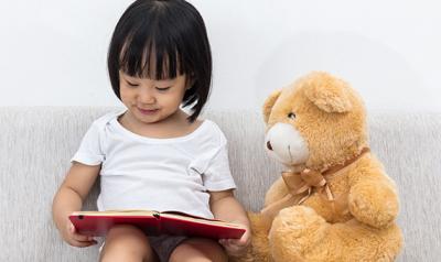 Young girl reading next to a teddy bear