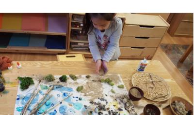 a child working on a craft project with grass, sticks, and sand