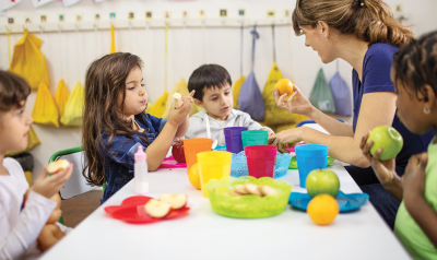 Children eat food at a table with their teacher.