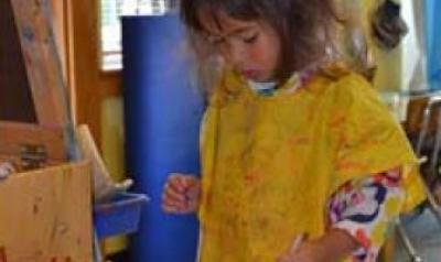 Child with paint smock on, standing near easel and paint containers.