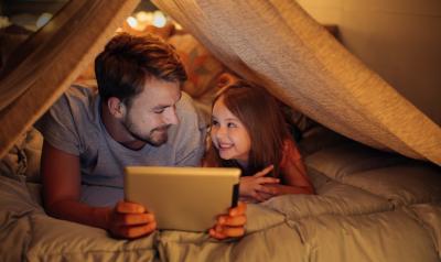 An adult reads to a child from a tablet screen under a tent indoors.