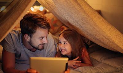 father and daughter playing on a tablet under a homemade fort. 