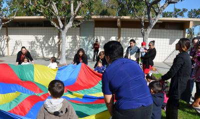A group of children and their parents play with a rainbow-colored parachute.
