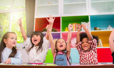 Diverse group of children in a classroom raising their hands in the air!