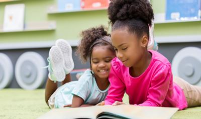 Two preschool girls on the floor in the classroom reading a book together