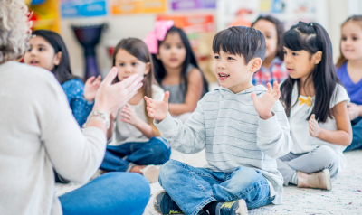 A teacher clapping with students in a circle on the floor.