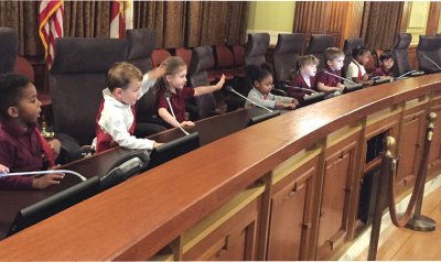 A group of preschool children sits at a city hall desk.