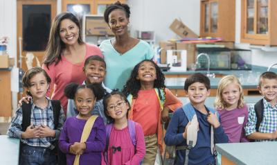 A Black and a white co-teacher stand with their group of young children in the classroom.