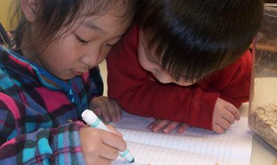 A young boy watching a young girl writing in a notebook