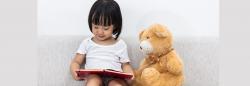 Young girl reading next to a teddy bear