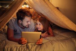 An adult reads to a child from a tablet screen under a tent indoors.