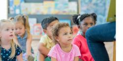 Children sit on the floor in a group as their teacher reads a story