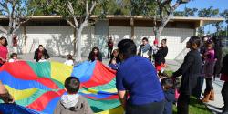 A group of children and their parents play with a rainbow-colored parachute.