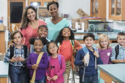 Two teachers and a group of young children standing and smiling in the classroom.