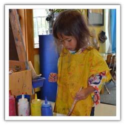 Child with paint smock on, standing near easel and paint containers.