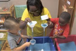 Teacher and children play with funnels at sand table.