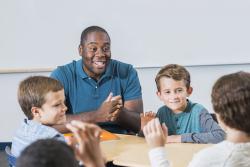 Young black male teacher of color sites at a table with group of white boys.
