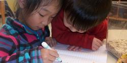 A young boy watching a young girl writing in a notebook