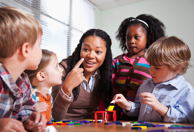 teacher sitting a table with a group of young students as they work on a project and she points to herself as if using herself as an example 